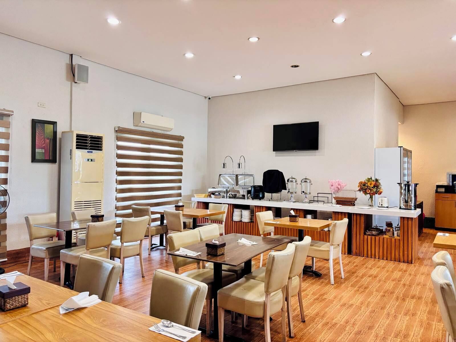 A modern hotel dining area with multiple tables and chairs, a kitchen counter with appliances, and a flat-screen TV mounted on the wall.