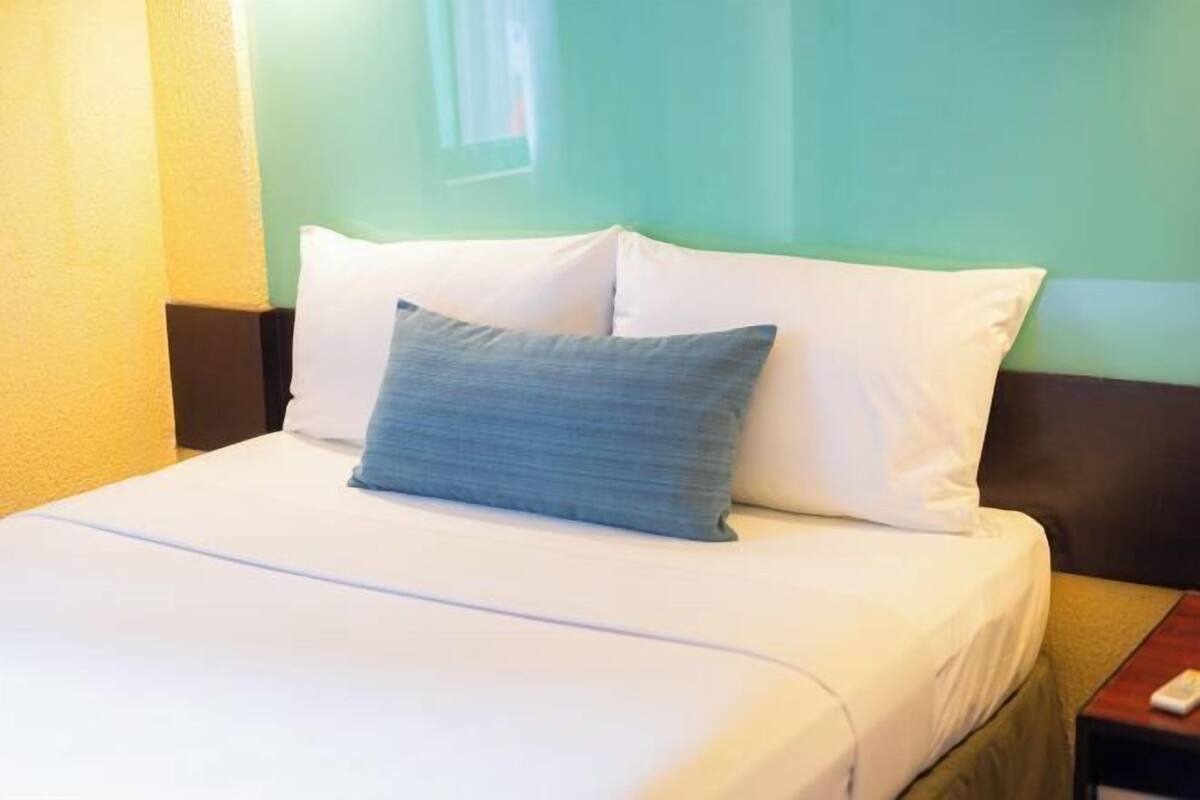 A neatly made bed with white bedding and a single blue pillow, a wooden nightstand with a remote control, and a green wall in the background.