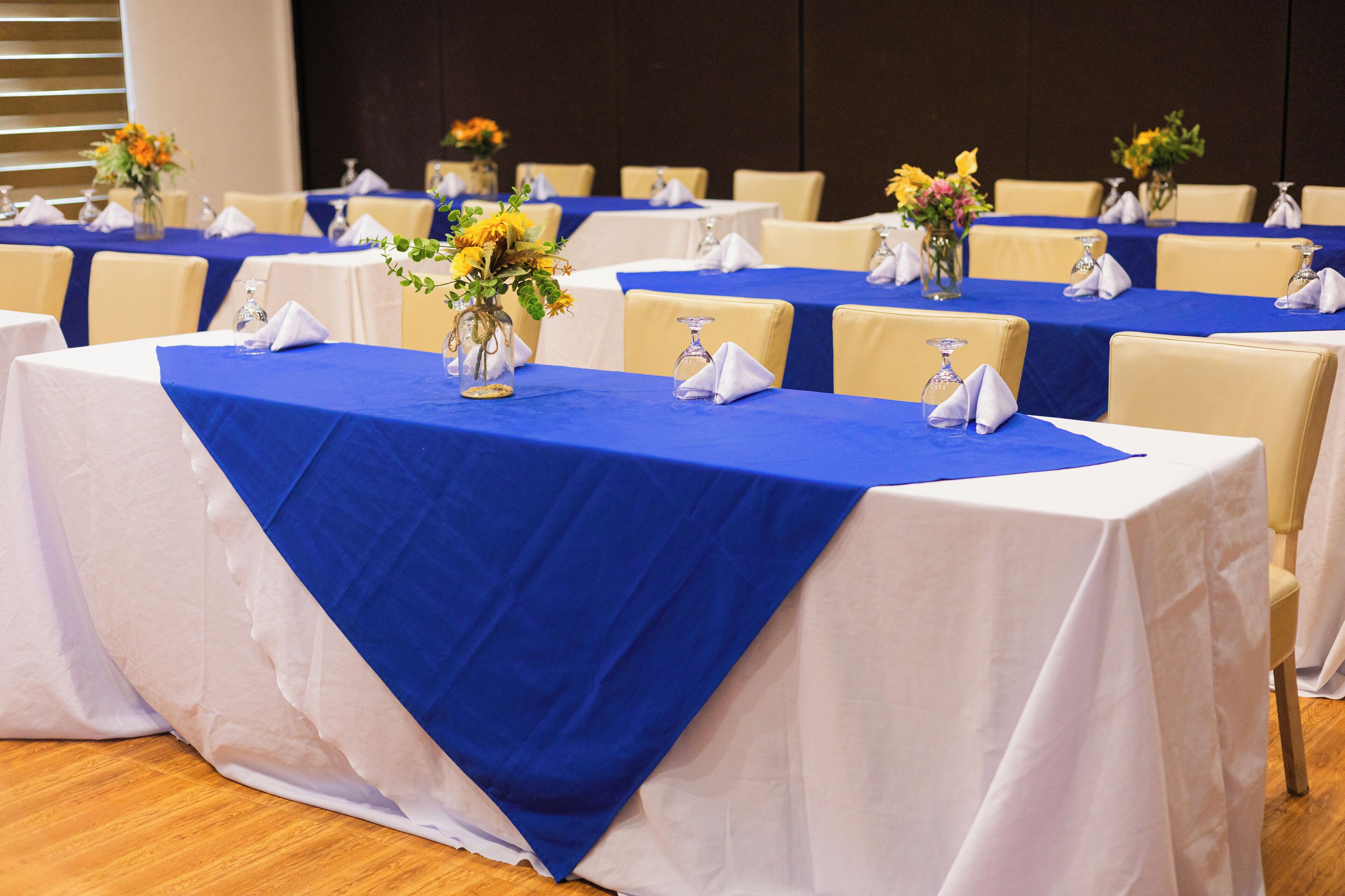 A conference room with tables set for a meeting. Each table has a blue tablecloth, white napkins, and a vase with flowers.