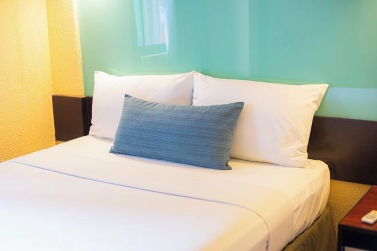 A neatly made bed with white bedding and a single blue pillow, a wooden nightstand with a remote control, and a green wall in the background.