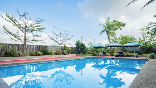 Une piscine à l’eau d’un bleu cristallin, entourée de chaises longues et de parasols, avec une clôture en bois et des plantes tropicales en arrière-plan.
