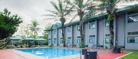 Una piscina con sillas de descanso y sombrillas, rodeada de palmeras y un edificio con varias ventanas.