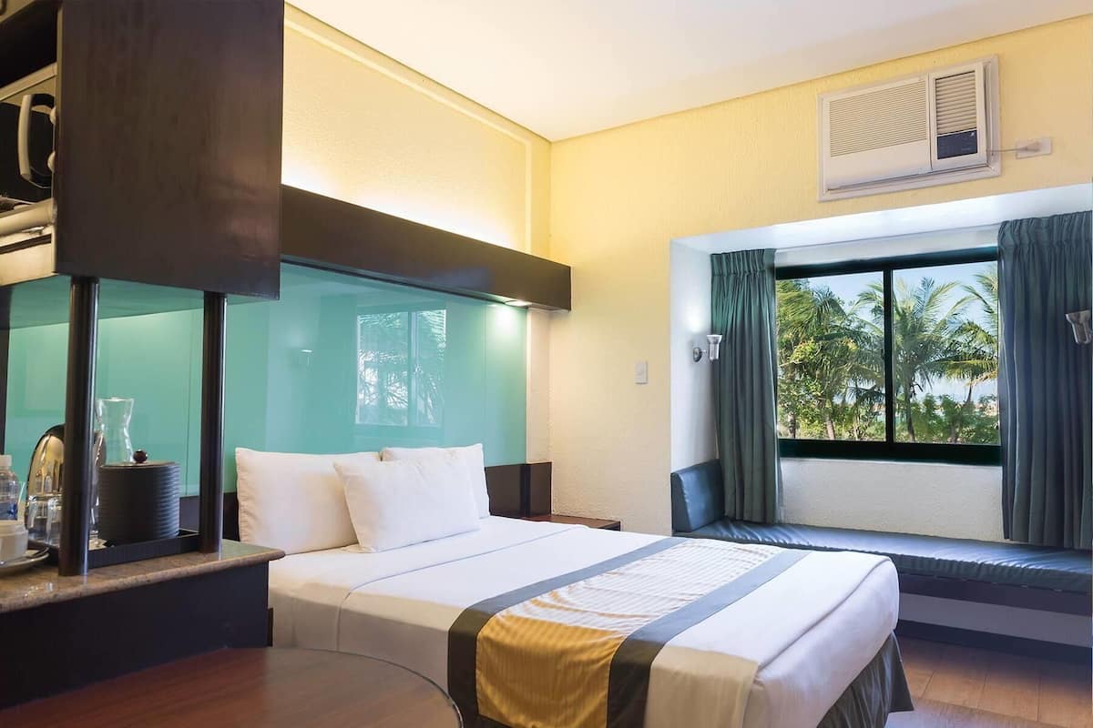 Ein Hotelzimmer mit Bett, Schreibtisch mit Lampe, Fenster mit Vorhängen und Klimaanlage.
