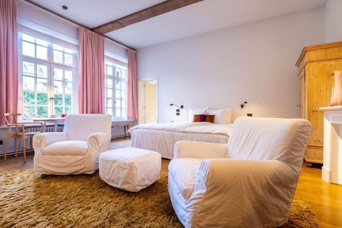 Una habitación con dos camas, una silla, una mesita y un ventanal con cortinas.