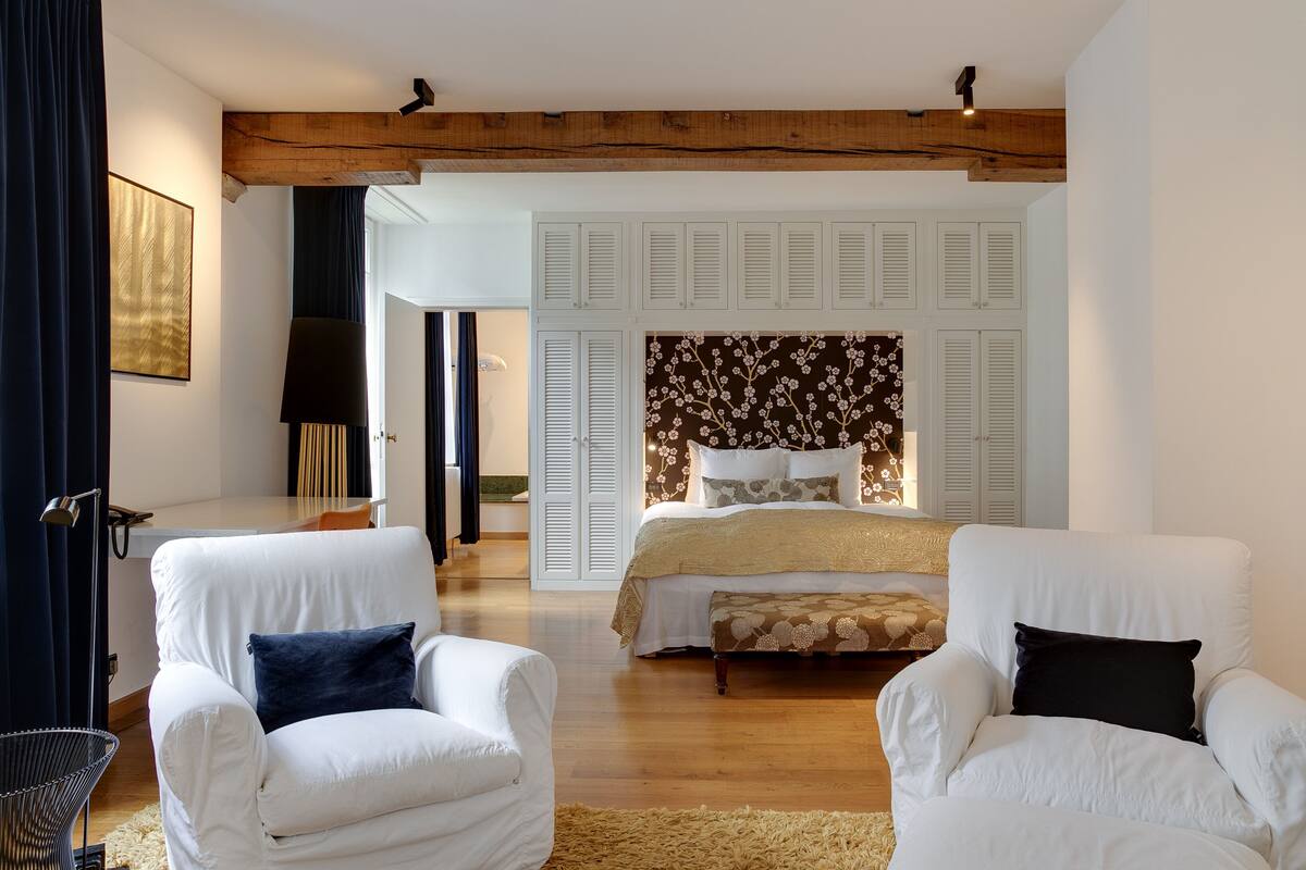 A bedroom with a large bed, two white armchairs, a wooden ceiling, and a floral headboard.