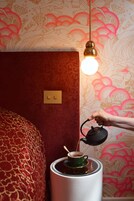A hand pouring tea from a teapot into a cup on a table, with a light bulb hanging above and a patterned wallpaper in the background.
