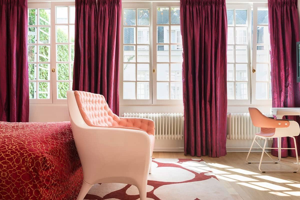 A bedroom with a bed, a chair, a radiator, and large windows with curtains.