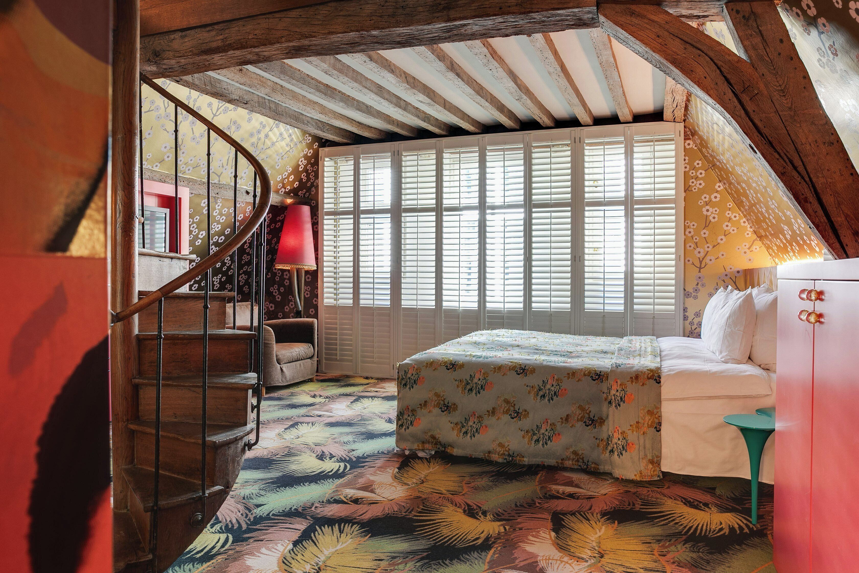 A bedroom with a wooden staircase, a bed with floral bedding, a window with shutters, and a red lamp.