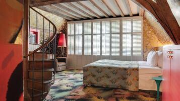 A bedroom with a wooden staircase, a bed with floral bedding, a window with shutters, and a red lamp.