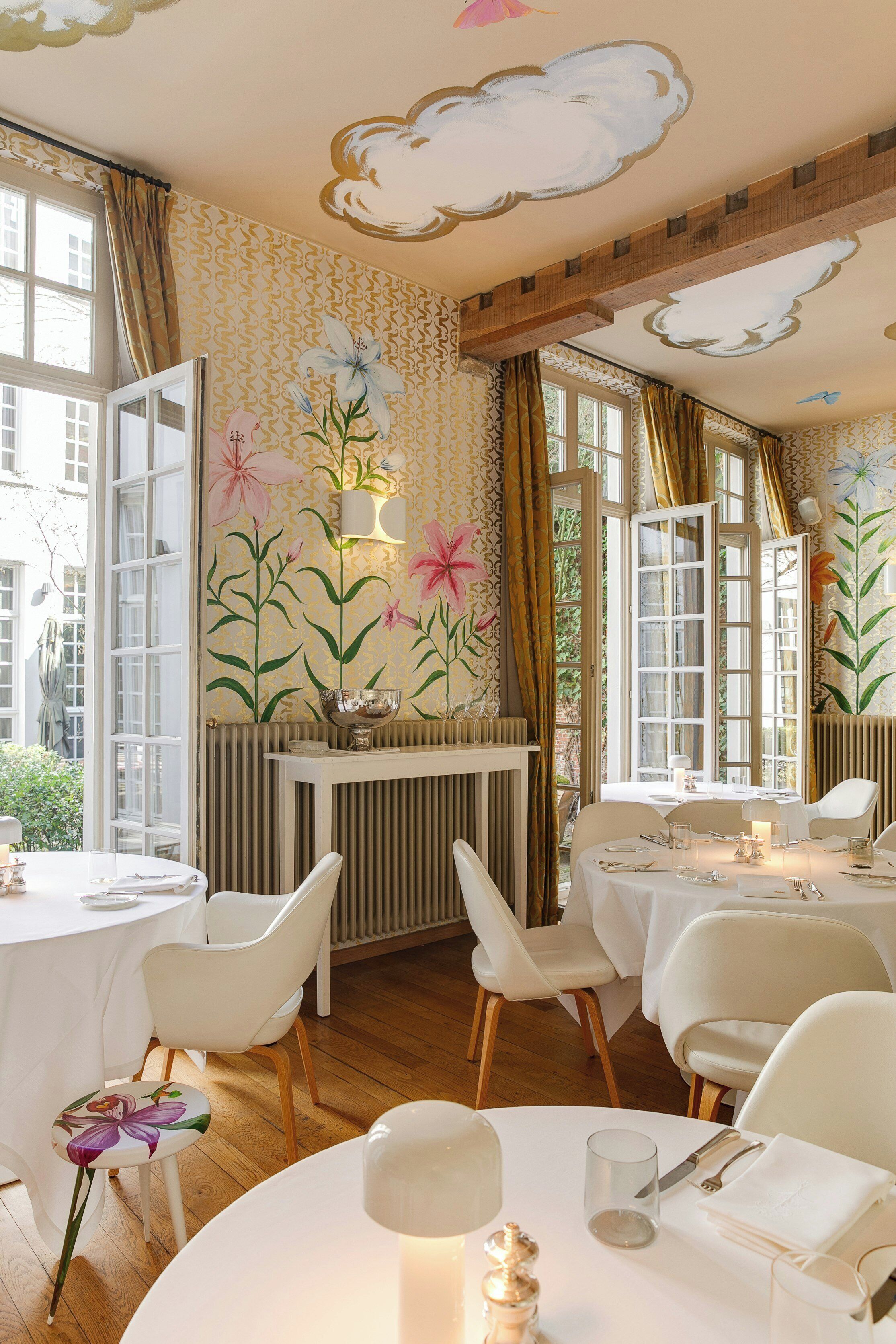 Comedor con sillas blancas, mesas redondas y decoración floral en la pared.