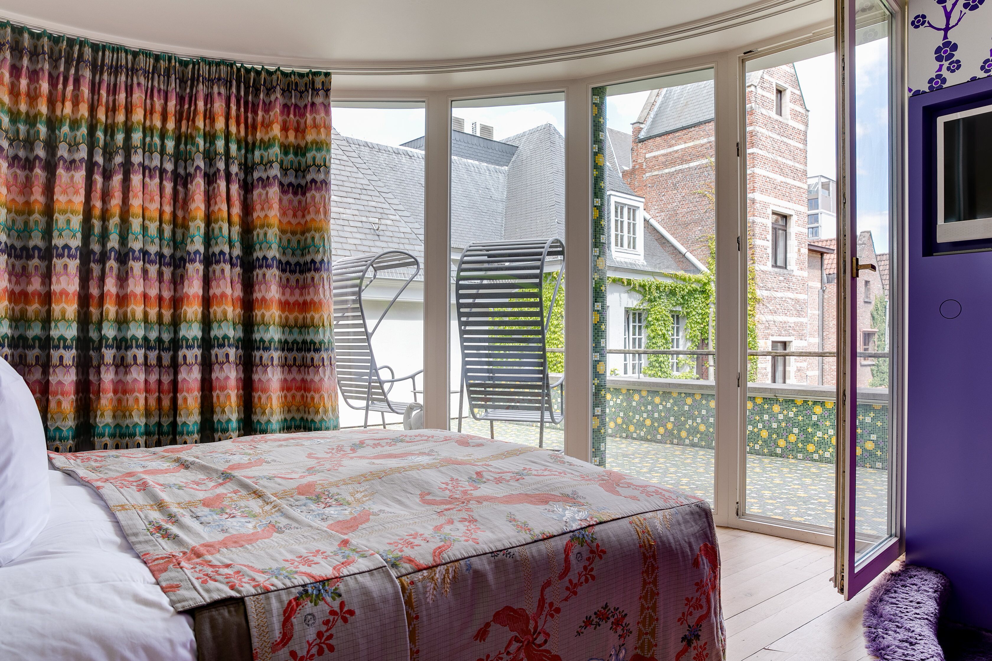 A bedroom with a bed, a colorful curtain, a window, and a view of a balcony with chairs.