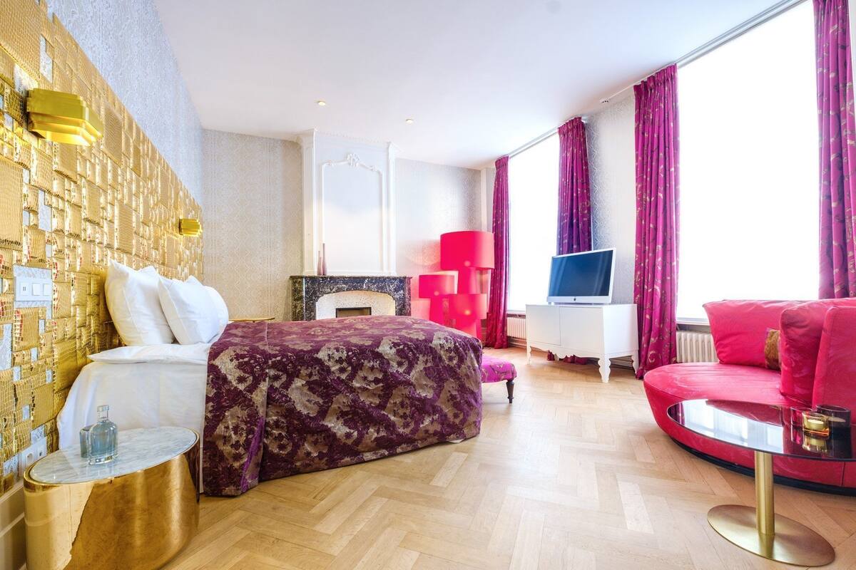 A hotel room with a bed, a TV, a red sofa, a small table, and a wall with a unique design.