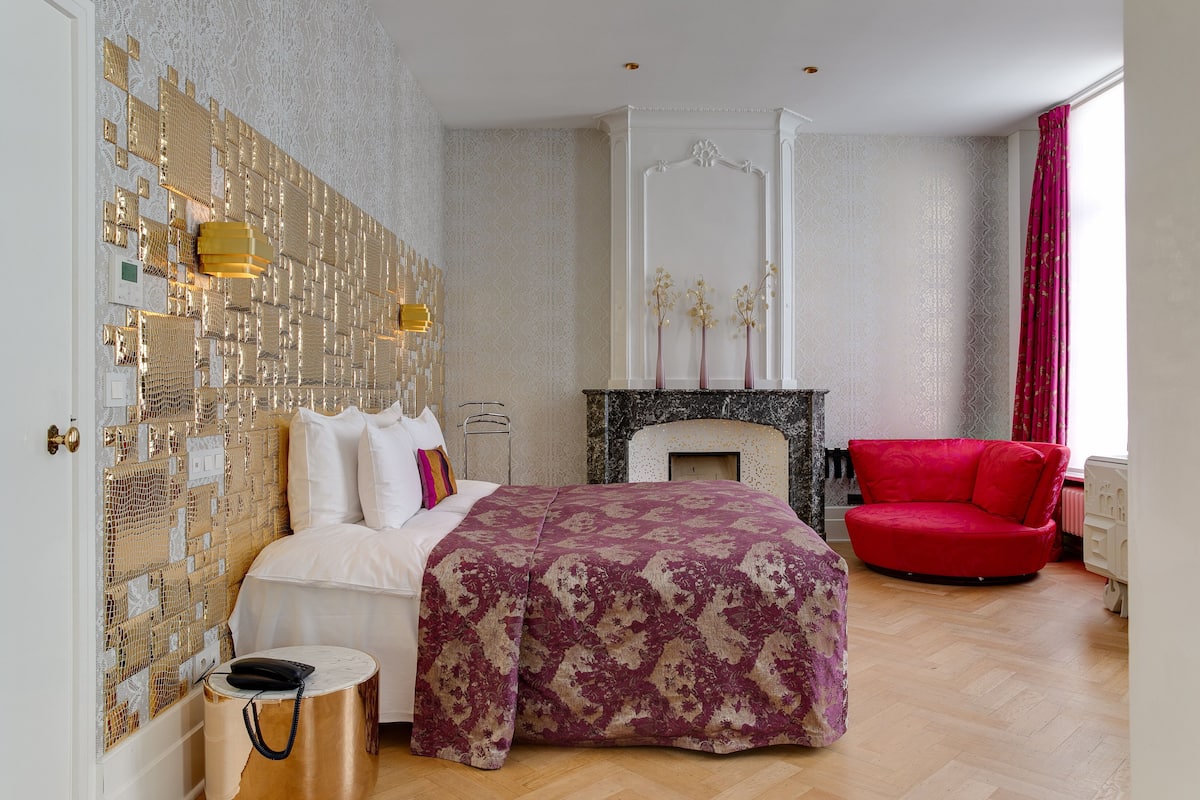 A bedroom with a patterned bedspread, a red armchair, a fireplace, and a decorative wall.