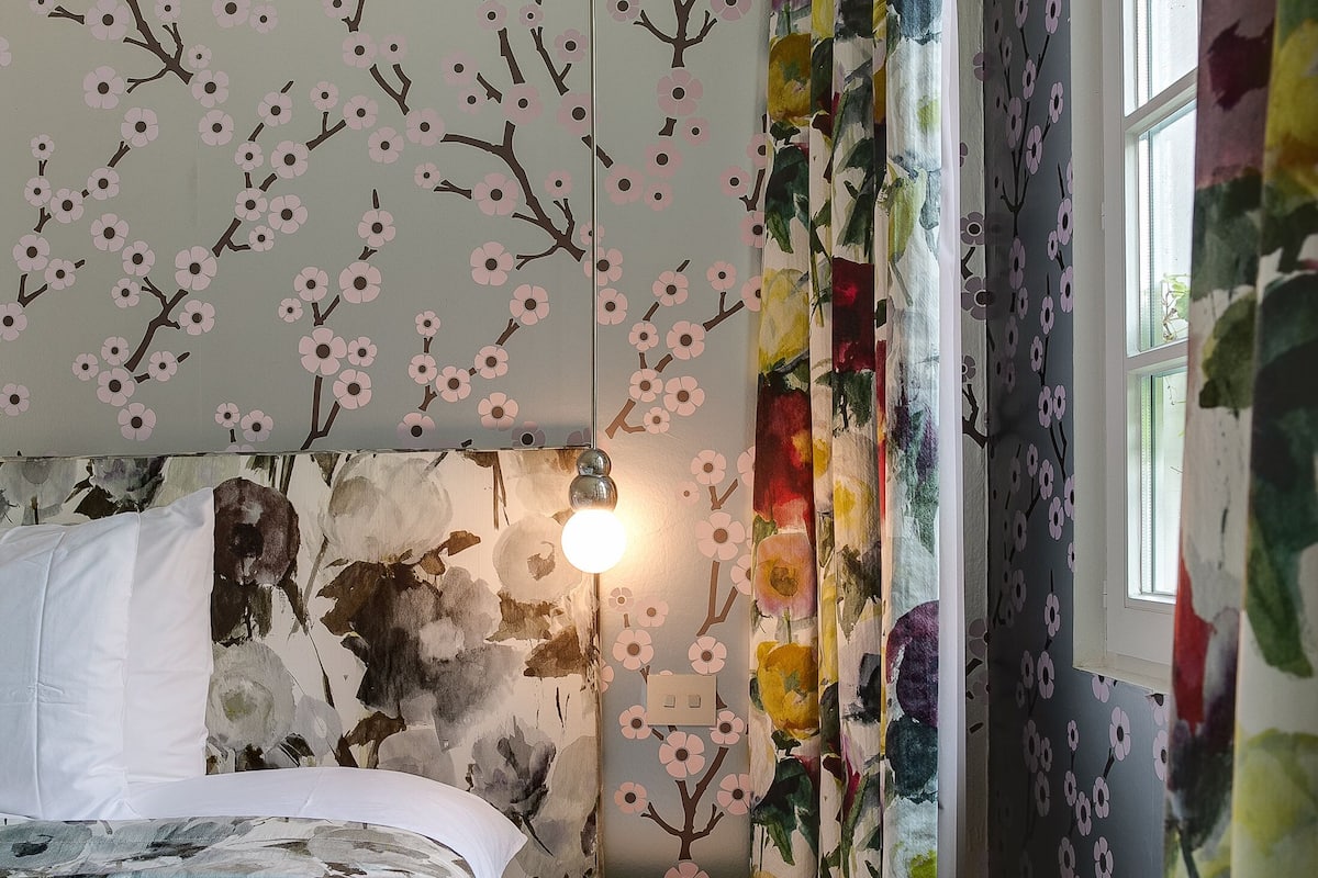 A bedroom with a floral wallpaper, a bed with a floral bedspread, a round blue ottoman, and a window with curtains.