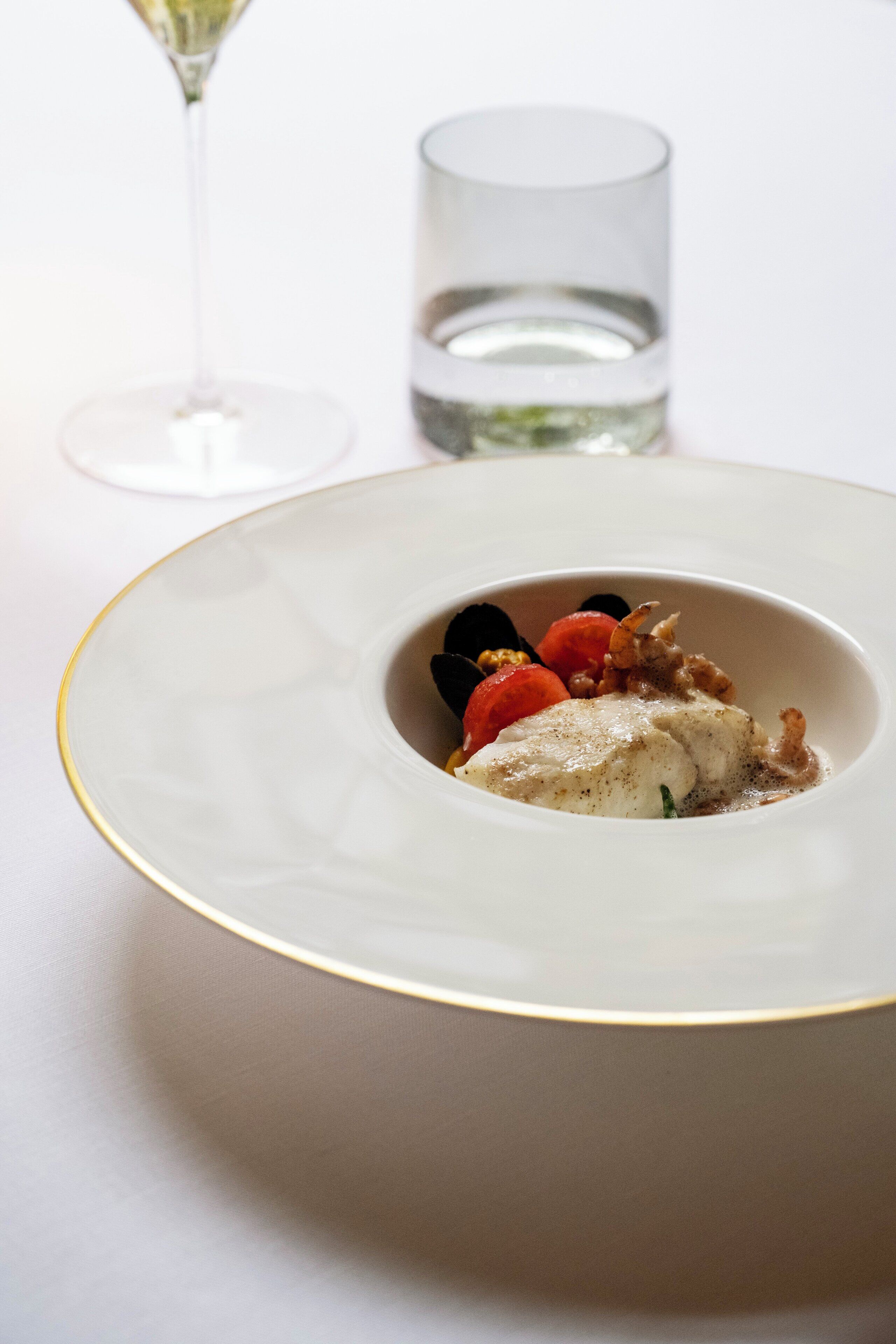 A white plate with a gold rim, containing a white fish dish, red tomatoes, and black olives.
