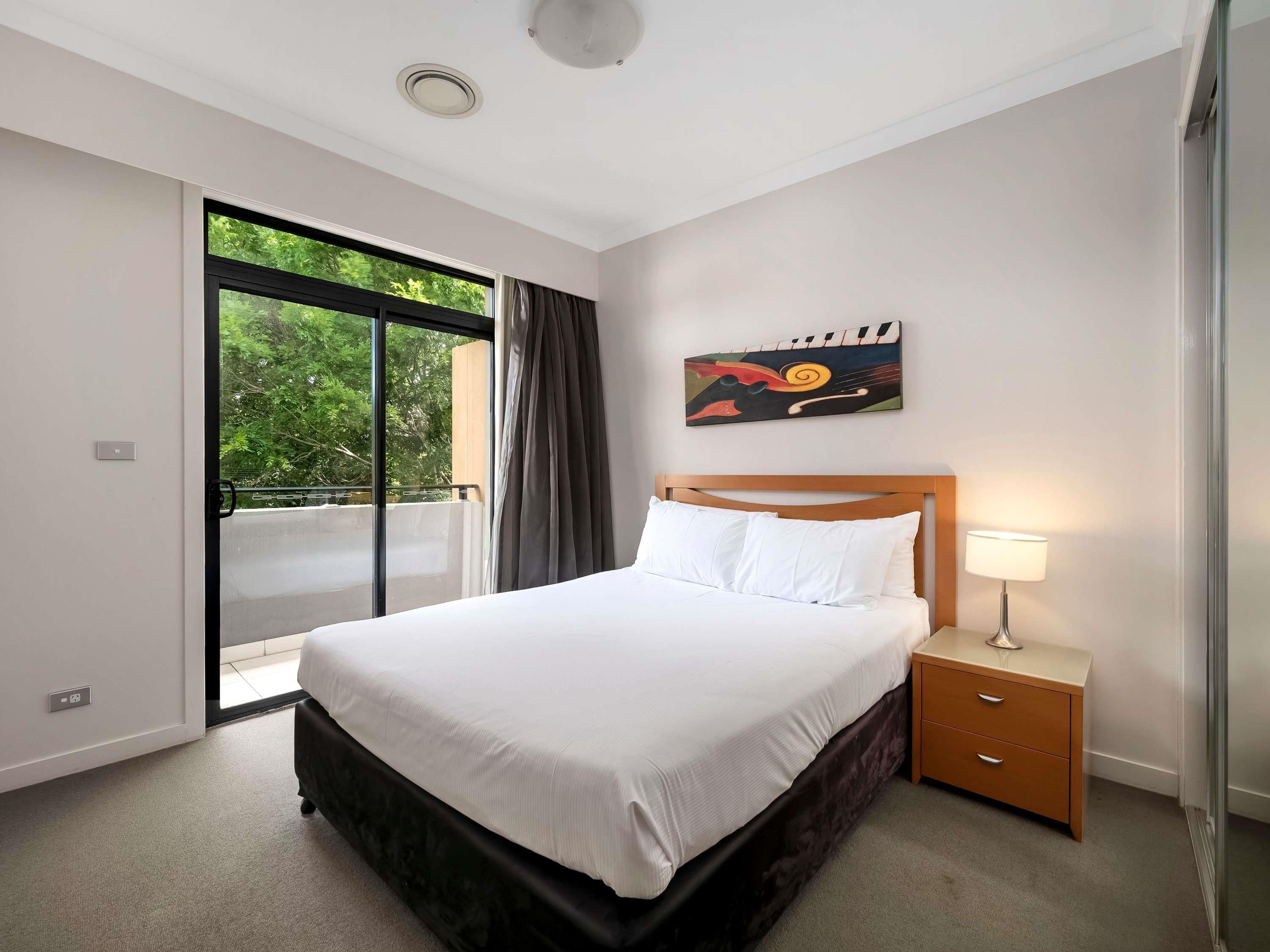 A hotel room with a bed, a nightstand, a lamp, a painting, and a sliding glass door leading to an outdoor area.