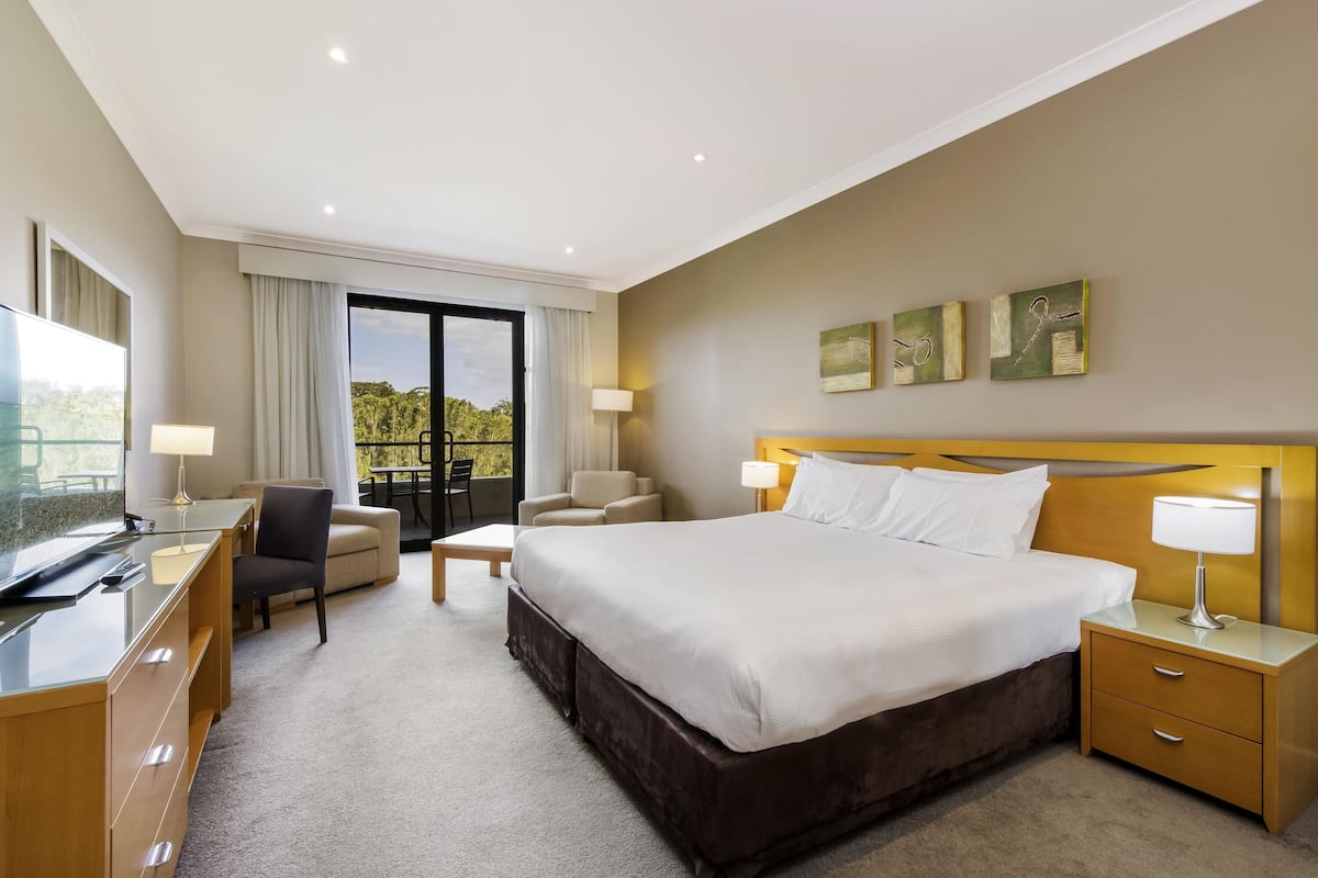 Habitación de hotel con cama, dos mesitas de noche, un escritorio con silla, un sofá, un balcón con mesa y sillas, y obras de arte en la pared.