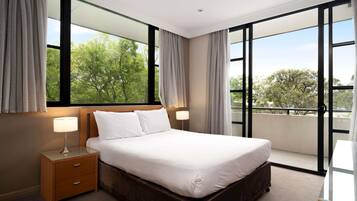 Hotelkamer met een groot bed, nachtkastje, lamp en uitzicht op bomen door het raam.