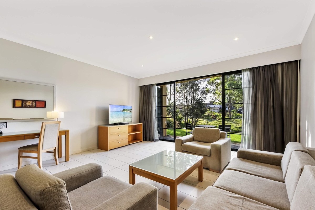 Una sala de estar moderna con un conjunto de sofá, una mesa de centro de vidrio, un televisor sobre un mueble de madera y vistas a un jardín a través de amplios ventanales.