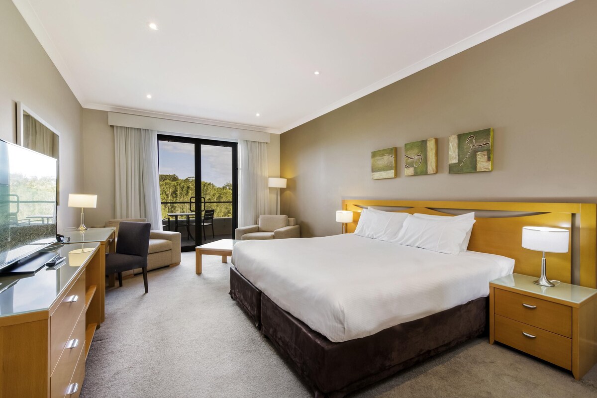 A hotel room with a bed, two bedside tables, a desk with a chair, a sofa, a balcony with a table and chairs, and artwork on the wall.