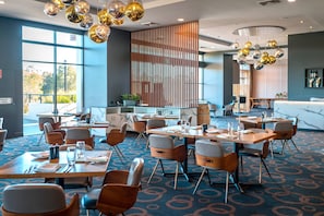 Een modern restaurant in een hotel, met meerdere tafels en stoelen, grote ramen en decoratieve, bolvormige lampen.