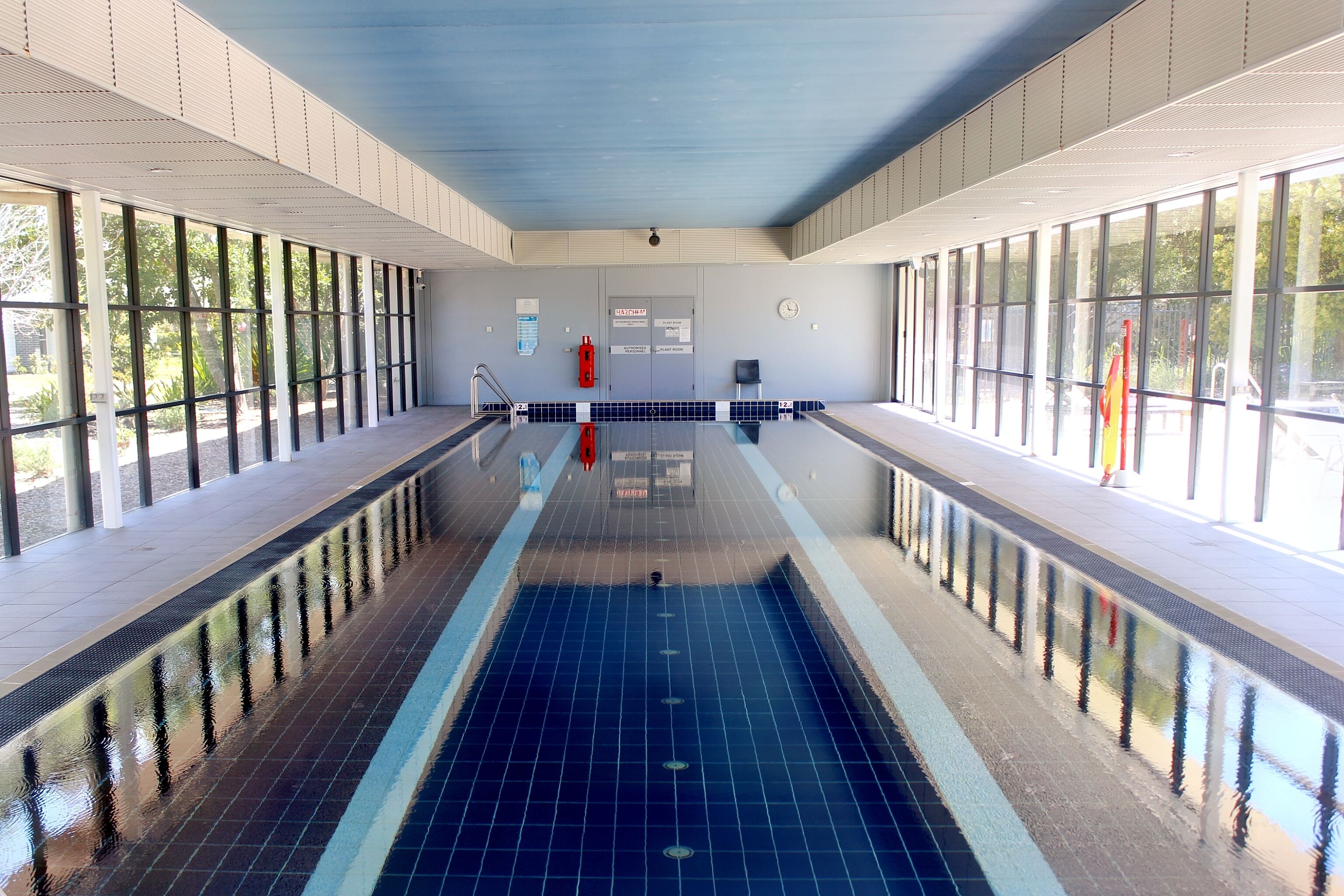 A large indoor swimming pool with clear glass walls, a tiled floor, and a visible exit door.