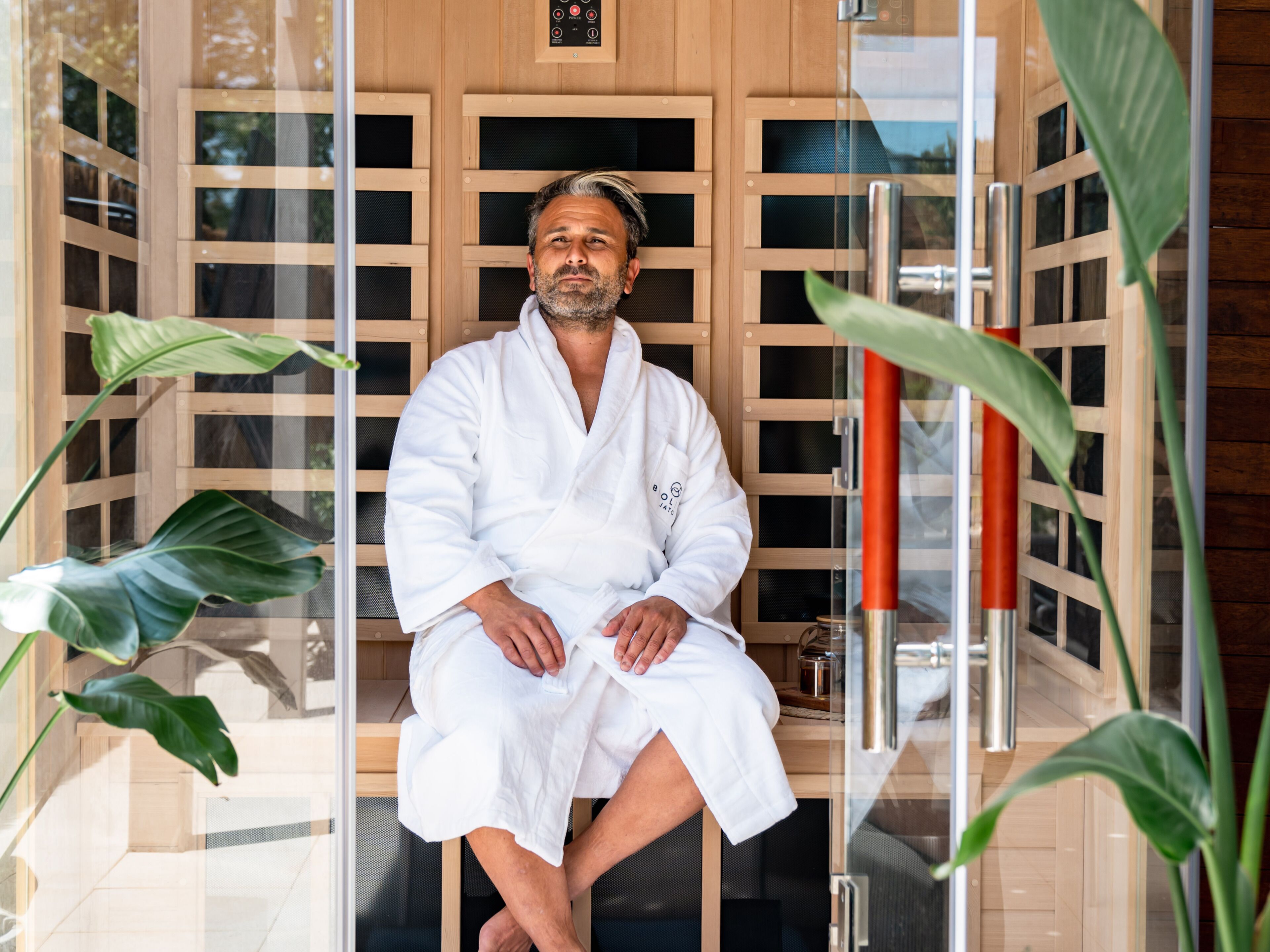 A person seated in a sauna room with wooden panels and glass doors, surrounded by large green plants.