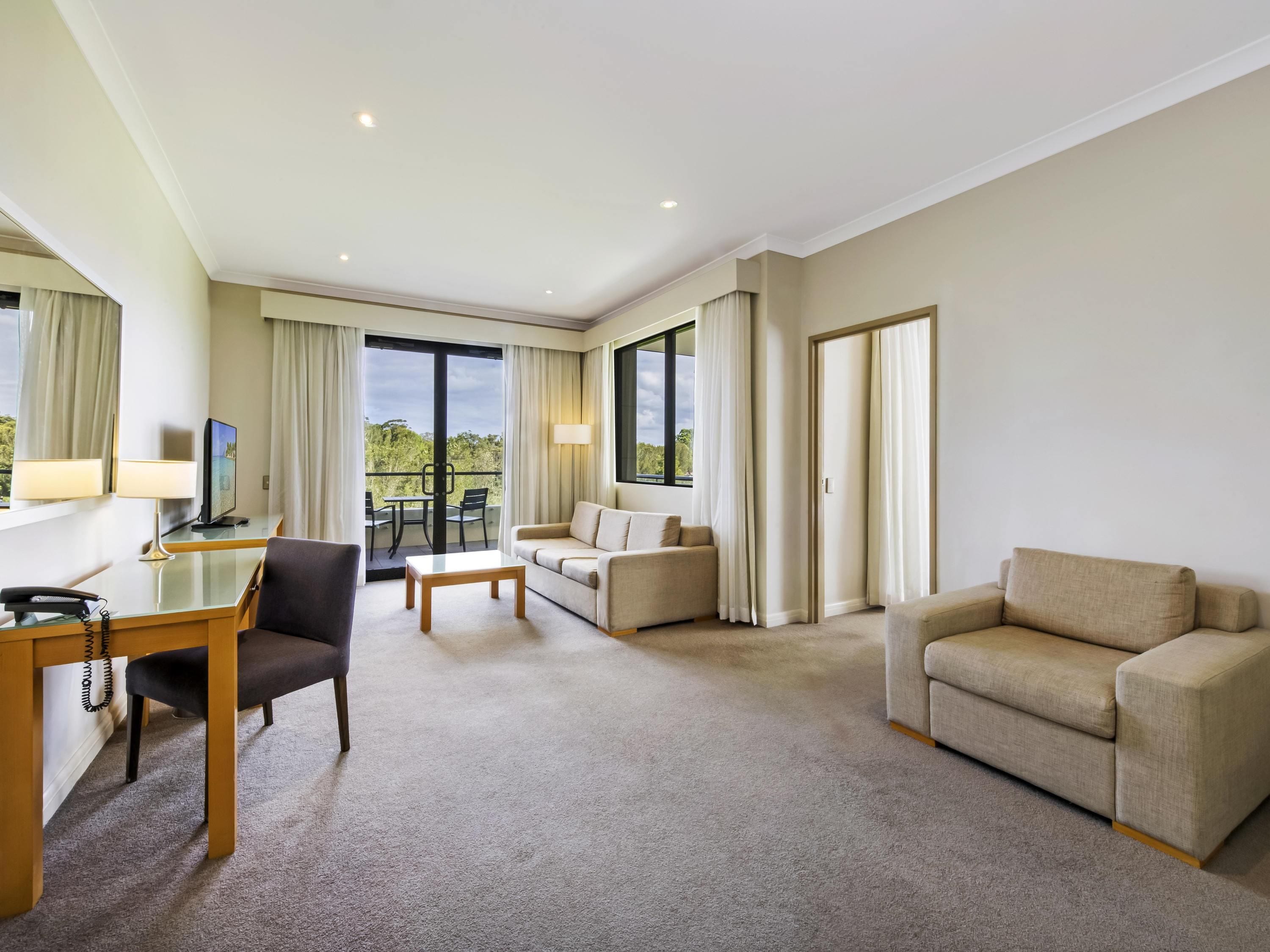 Habitación de hotel con escritorio, dos sofás, una silla, un balcón con mesa y televisión.