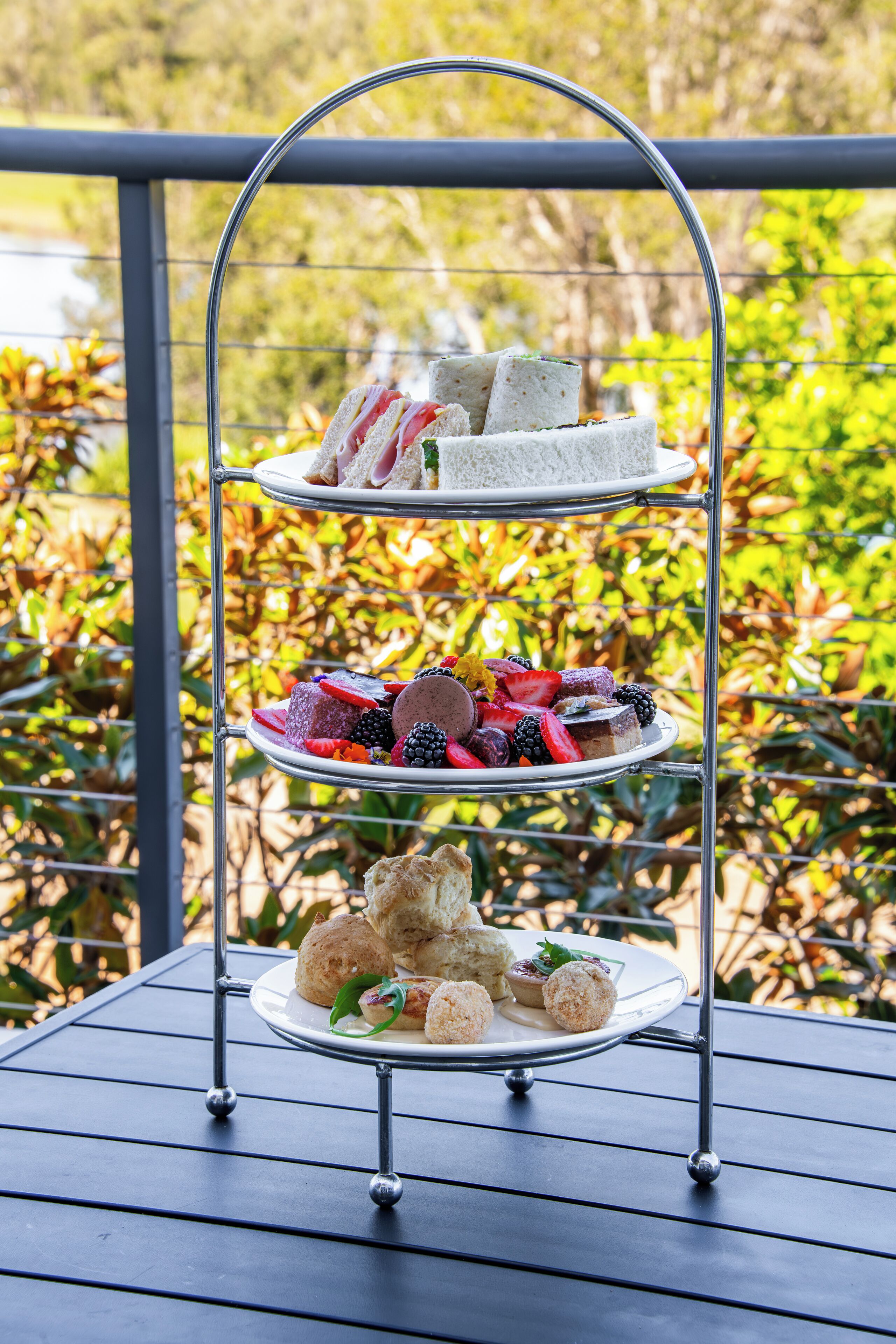 A three-tiered outdoor dessert stand with various pastries and sandwiches on a metal table.