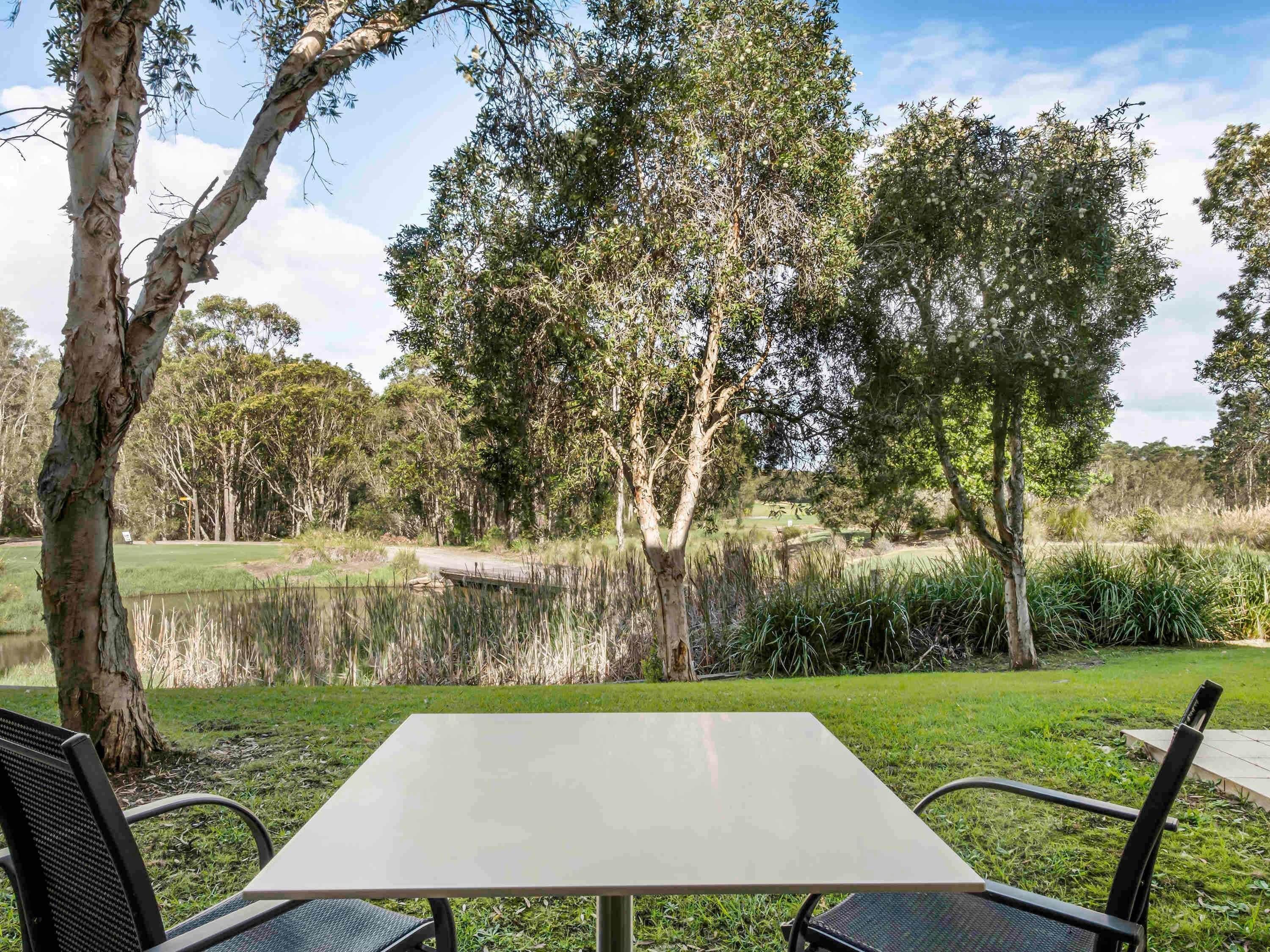 A table and chairs in a park with trees and a pond.