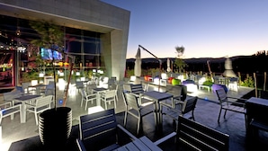 An outdoor patio area with tables and chairs, a modern building in the background, and a clear sky.