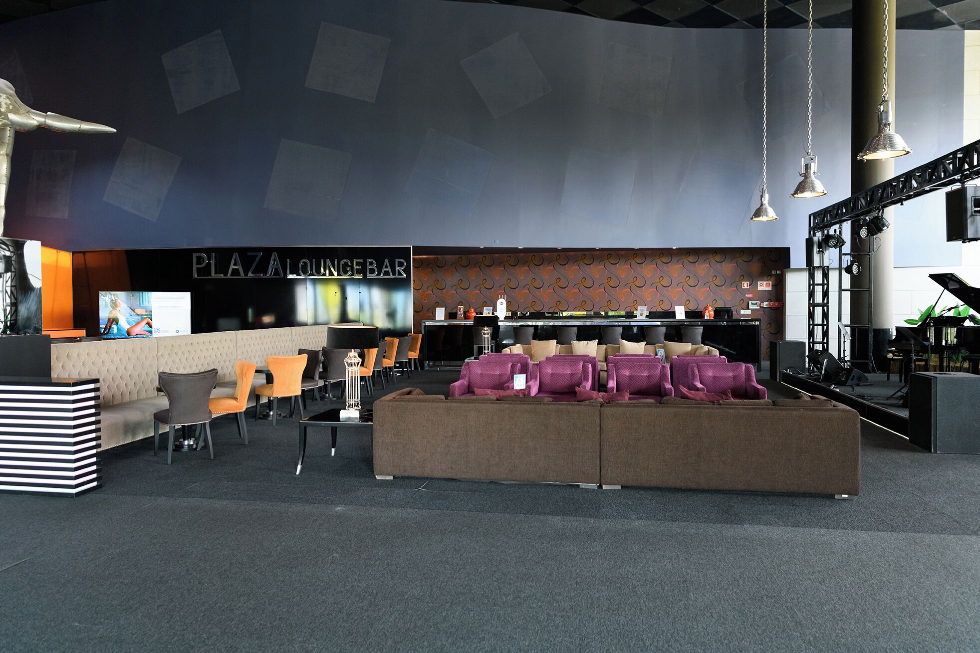 Uma área de estar com assentos, um piano e um bar chamado 'PLAZA LOUNGEBAR'.