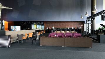 Uma área de estar com assentos, um piano e um bar chamado 'PLAZA LOUNGEBAR'.