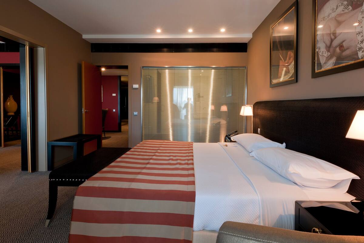 A hotel room with a bed, bedside table, lamp, and a large window with blinds.
