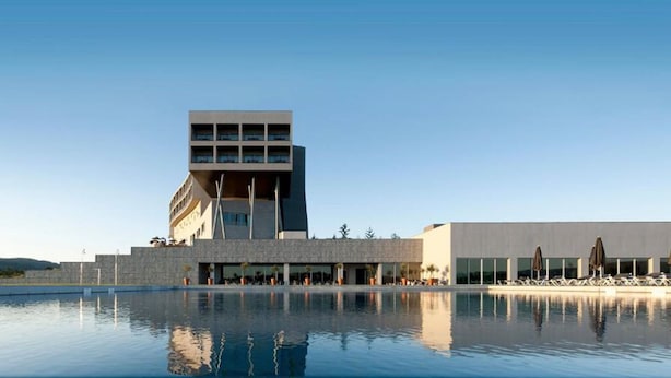 A modern hotel with a pool in front, featuring a unique architectural design with a prominent tower and a flat-roofed section.