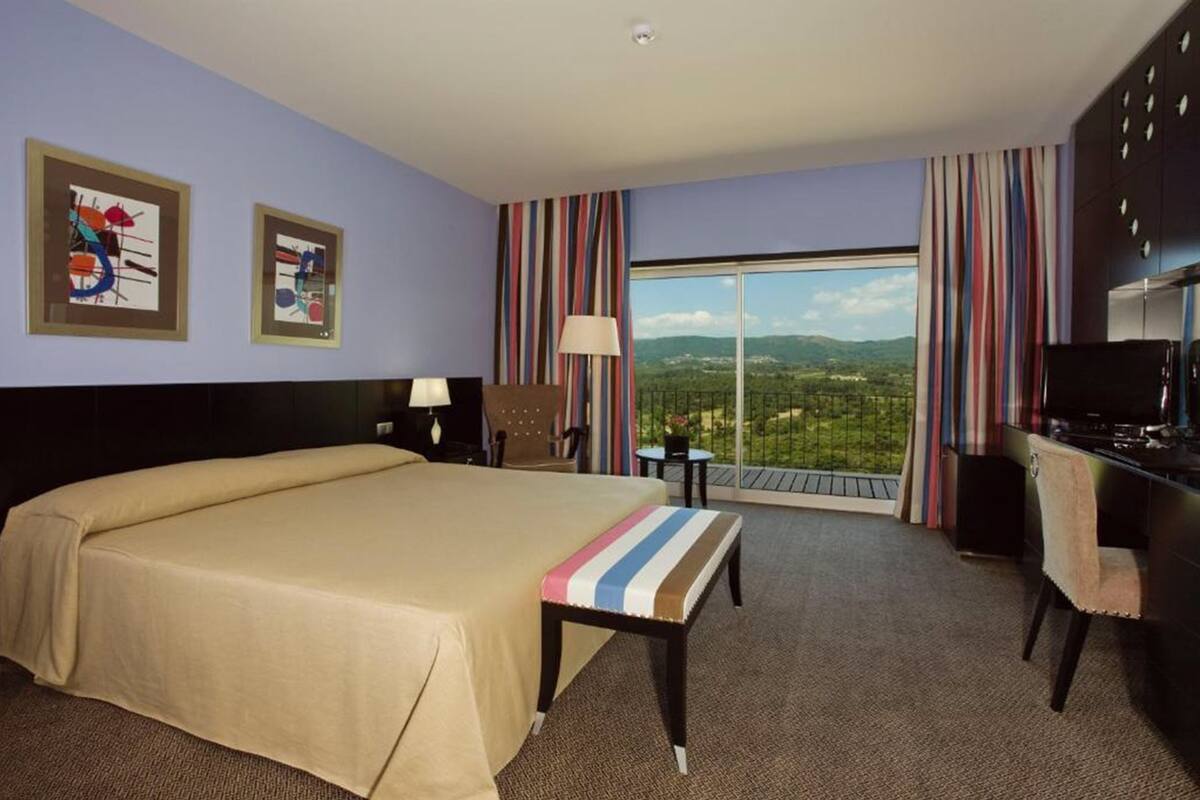 Habitació d'hotel amb un llit gran, escriptori, cadira, làmpada i vista al paisatge.