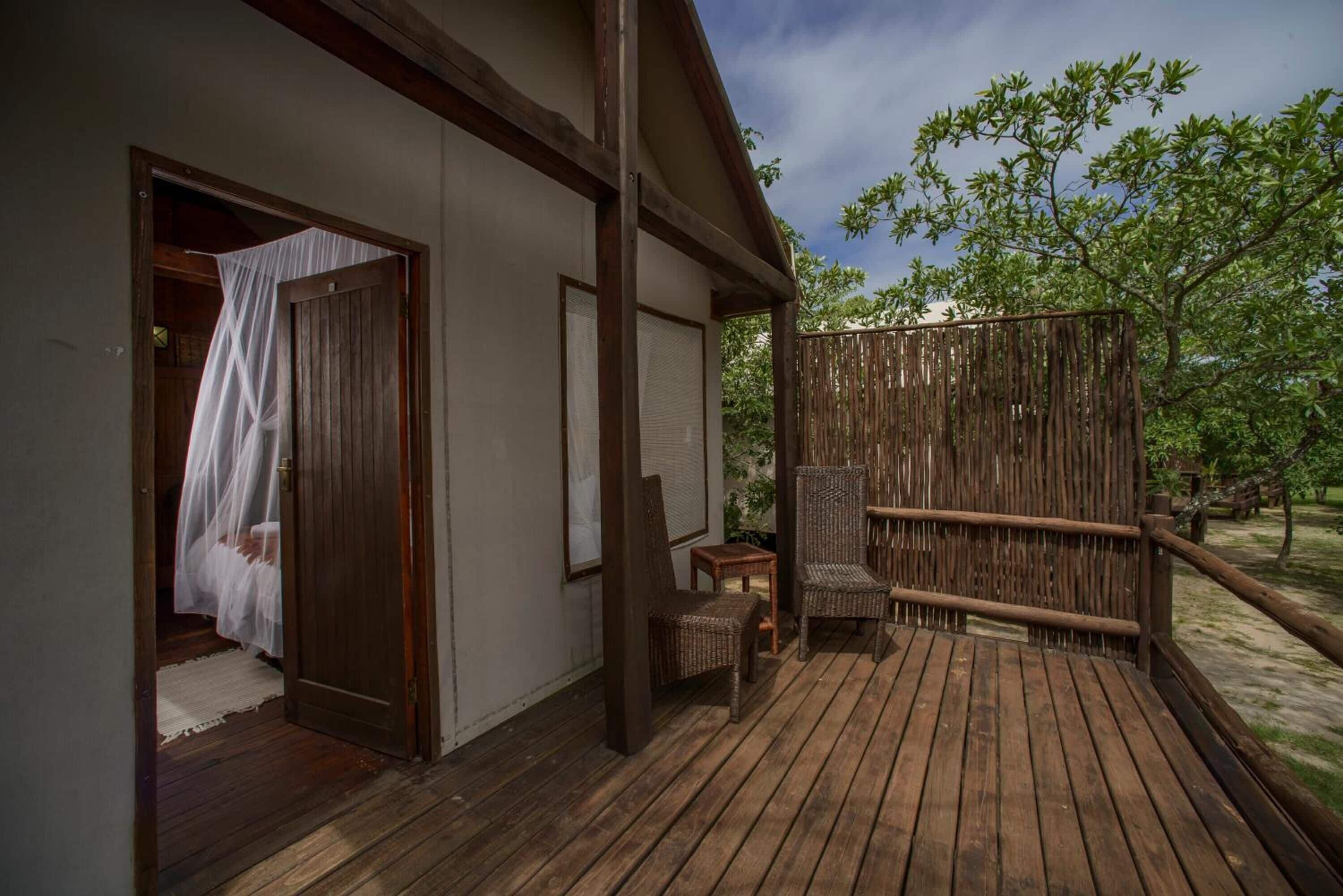 Una terraza de madera con sillas de mimbre, una valla de madera y una puerta que da a una habitación con cama.