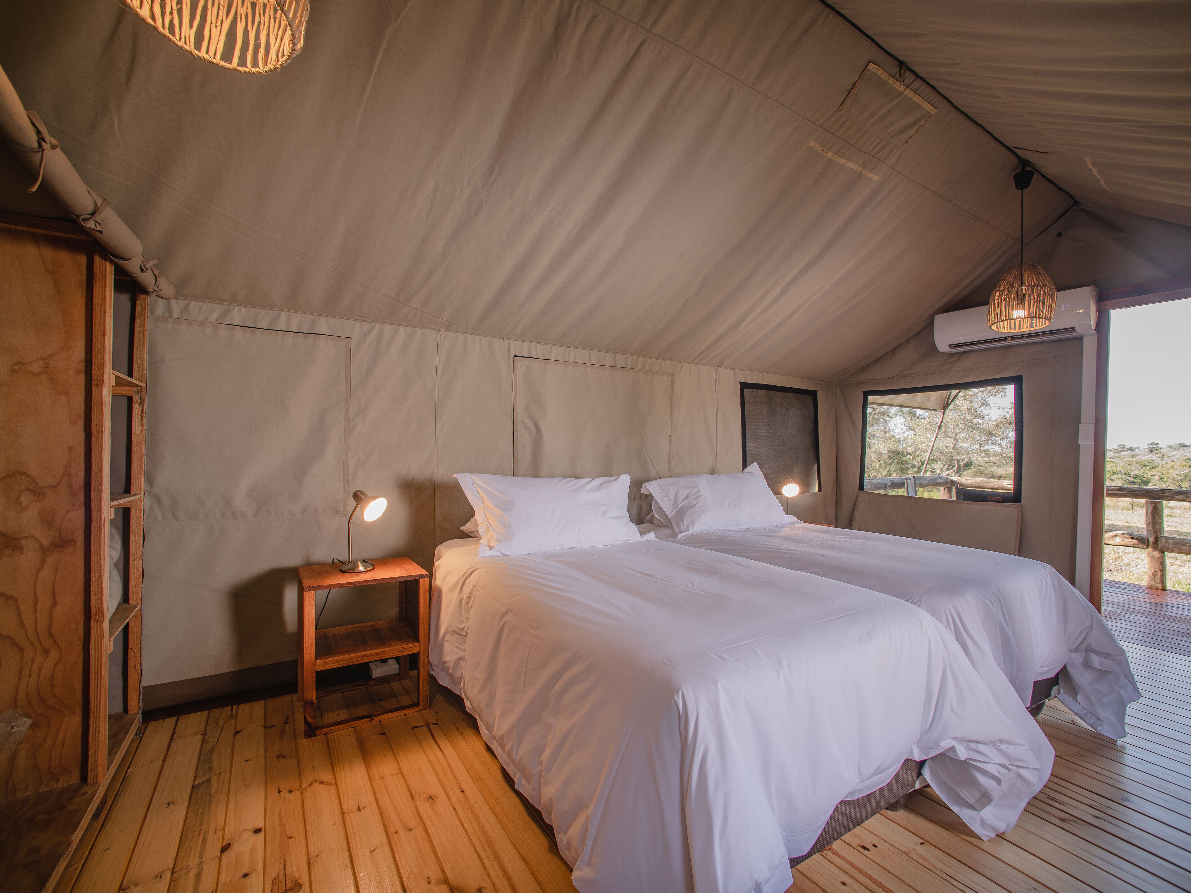 A tent-style room with two beds, a wooden floor, and a small table with a lamp.