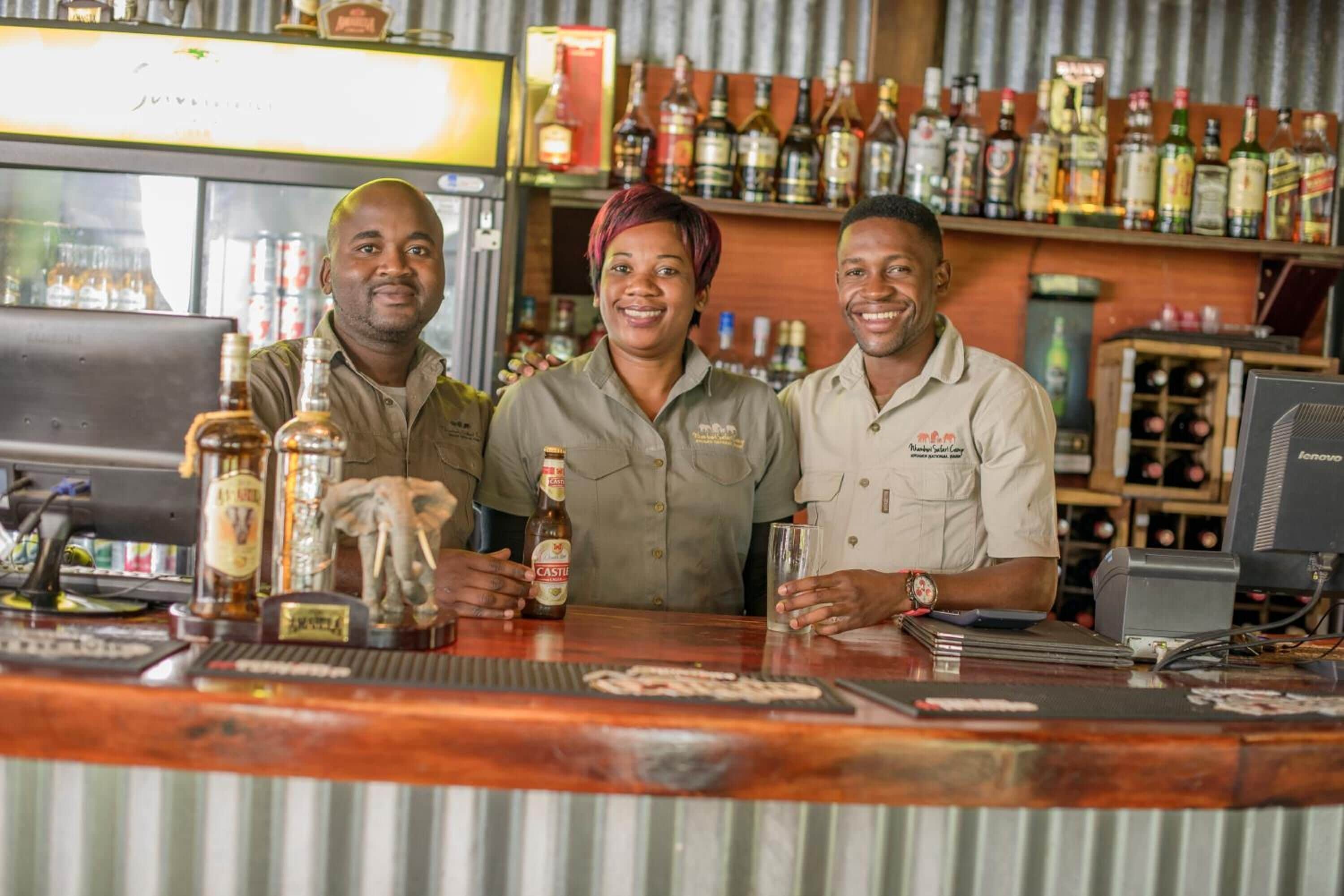 Three people standing behind a bar counter with various bottles and a computer monitor.