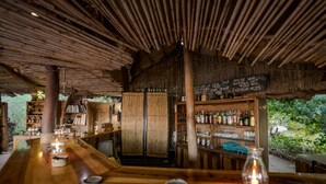 An indoor bar area with wooden furniture, a bamboo ceiling, and shelves stocked with bottles and glasses. - Taita Falcon Lodge (Livingstone)
