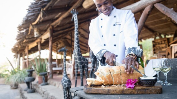 A chef slicing bread on a wooden board with a bowl of butter and two glasses of white wine on the side. - Taita Falcon Lodge (Livingstone)