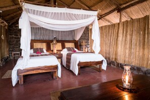 A room with two wooden beds under a canopy, a stone wall, and a wooden table with a candle. - Taita Falcon Lodge (Livingstone)