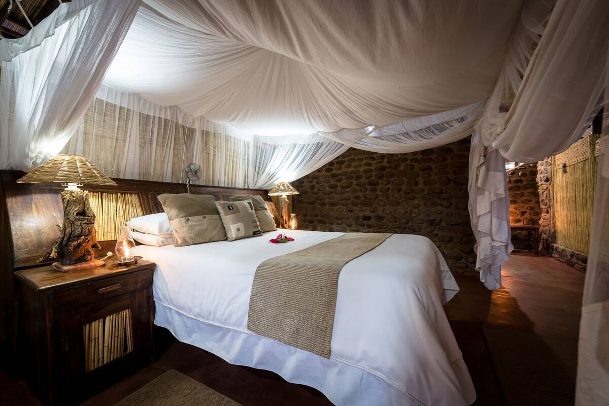 A bed with white linens, a wooden nightstand, a bedside lamp, and a stone wall.