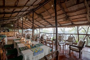 A rustic dining area with wooden tables and chairs, a thatched roof, and a view of trees. - Taita Falcon Lodge (Livingstone)