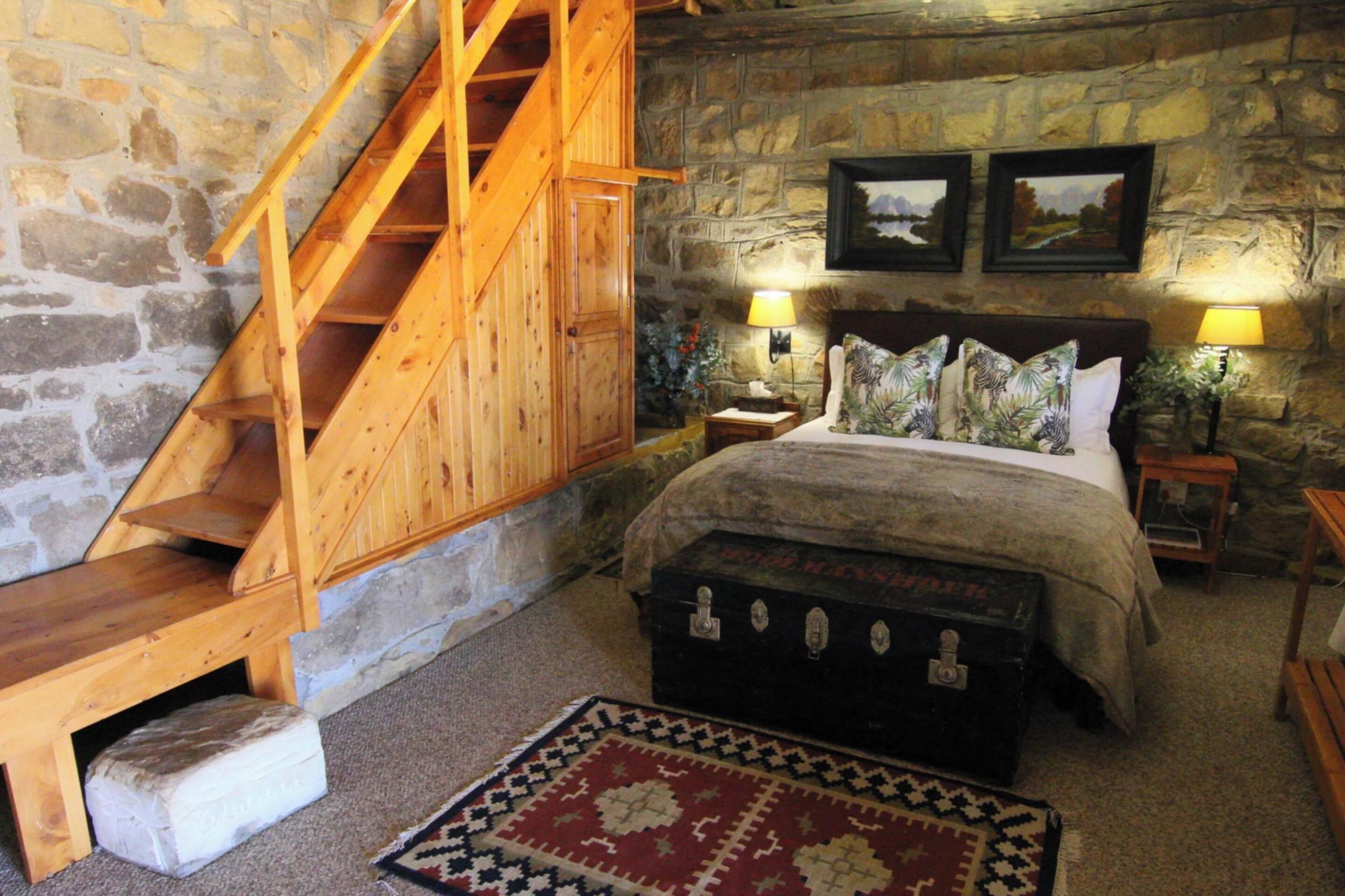 Un dormitorio con pared de piedra, escalera de madera, una cama con ropa de cama estampada, dos lámparas de noche y una alfombra decorativa.