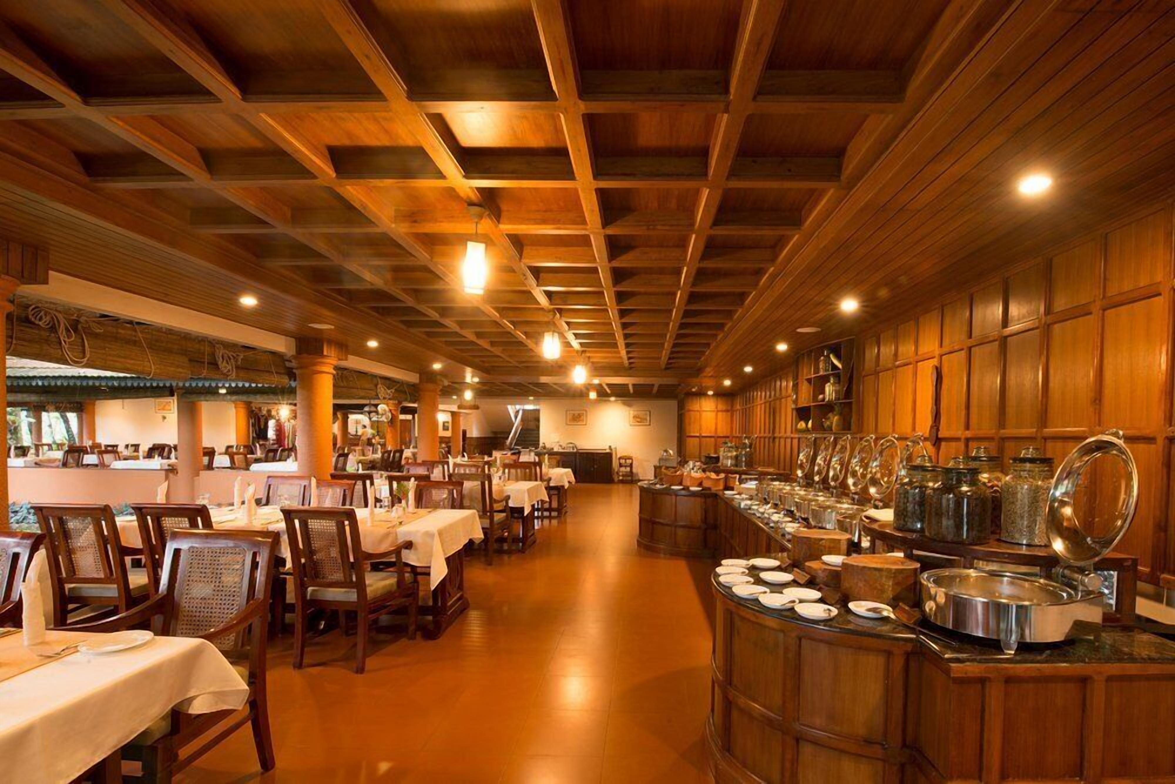 A restaurant with wooden ceiling beams, tables with white tablecloths, and a buffet area with food containers.