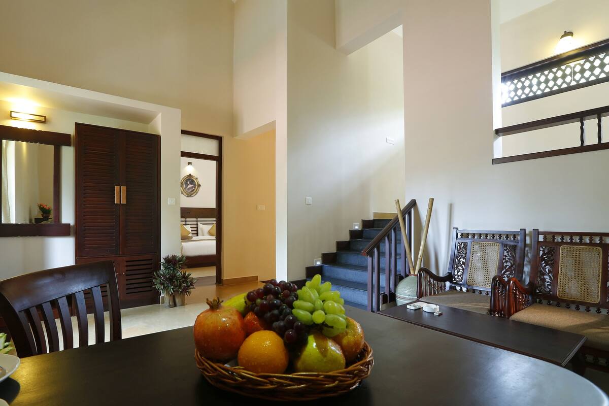 Un comedor con una mesa dispuesta para dos personas, una cesta de mimbre con fruta y una escalera que sube a un nivel superior.