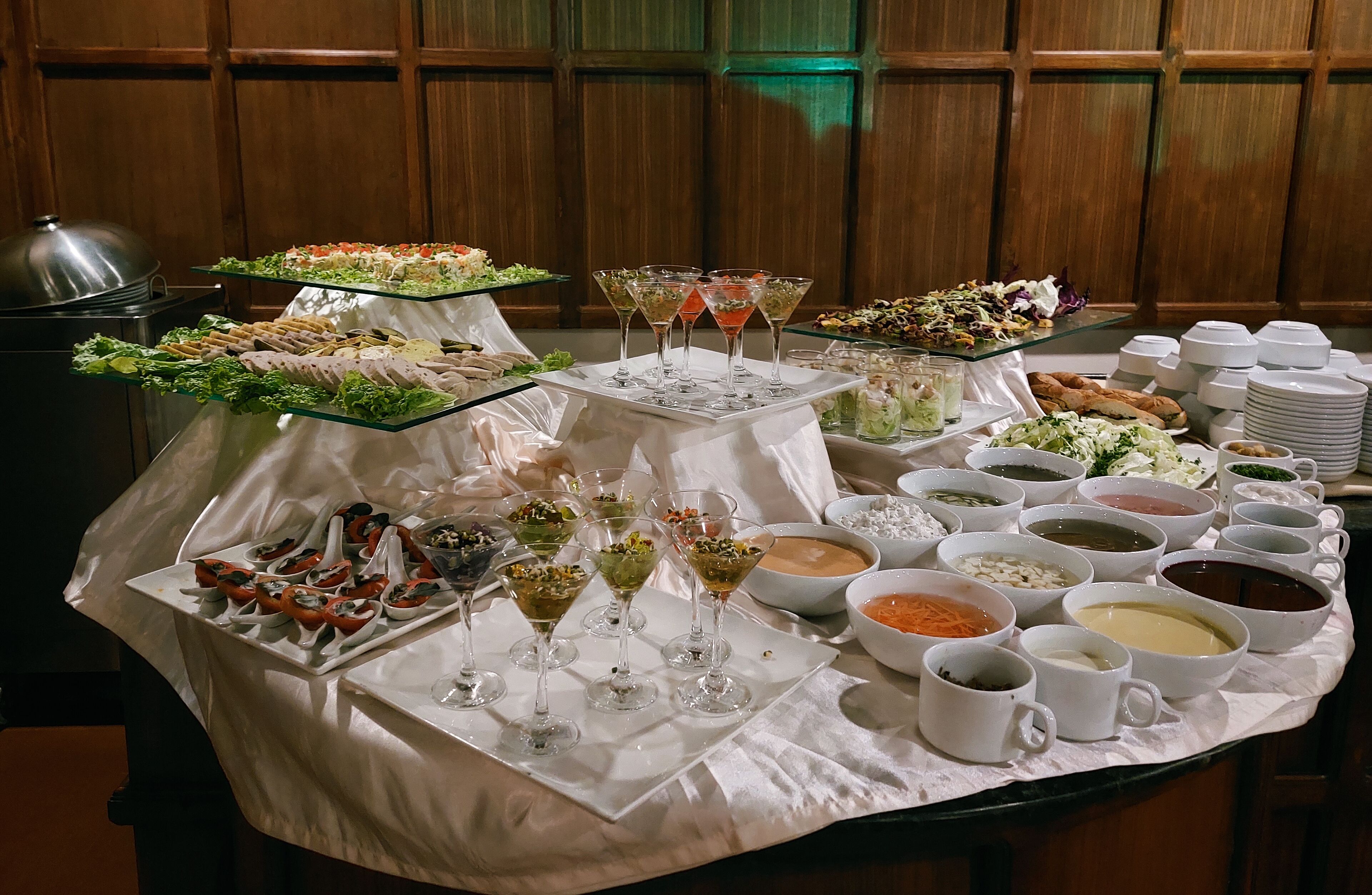 A buffet table with various food items and drinks, including salads, desserts, and beverages, set against a wooden paneled wall.
