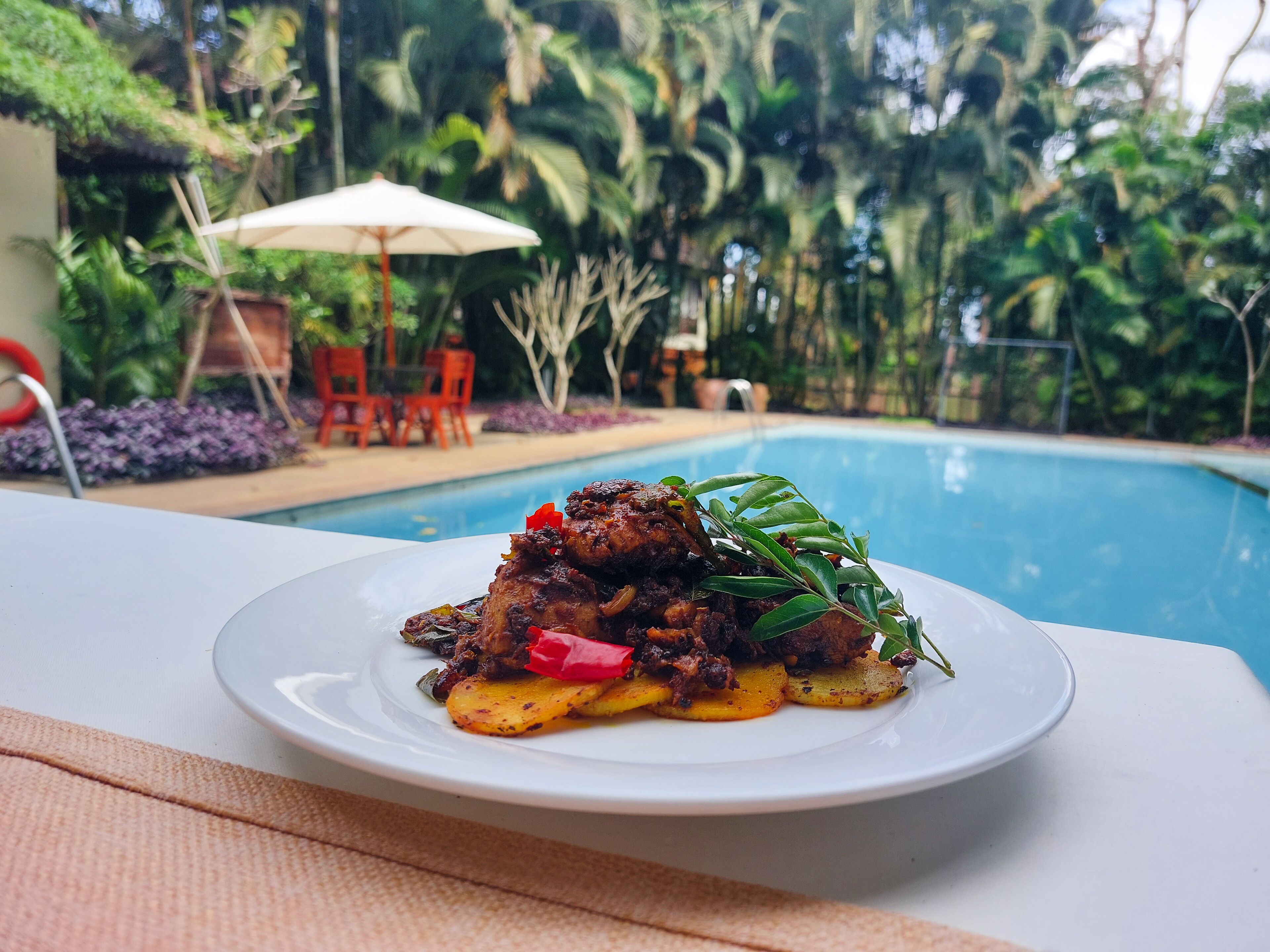 Una mesa de comedor con un plato de comida, una piscina y un jardín tropical al fondo.