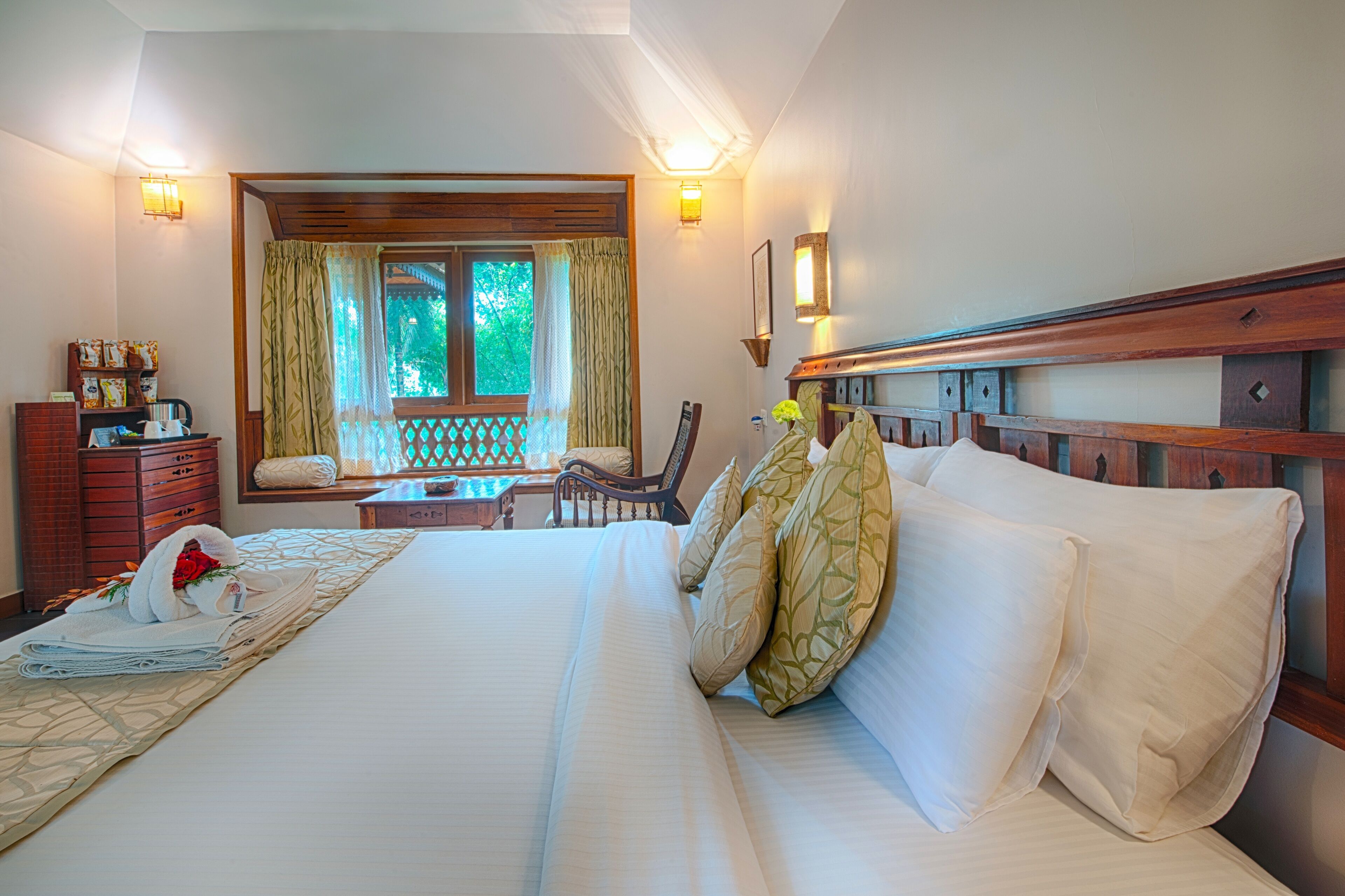 A hotel room with a large bed, a wooden headboard, a desk with a chair, a window with curtains, and wall-mounted lights.